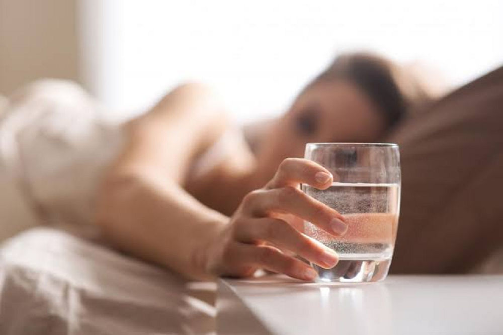 Mulher segurando copo de água, deitada na cama, o copo em primeiro plano