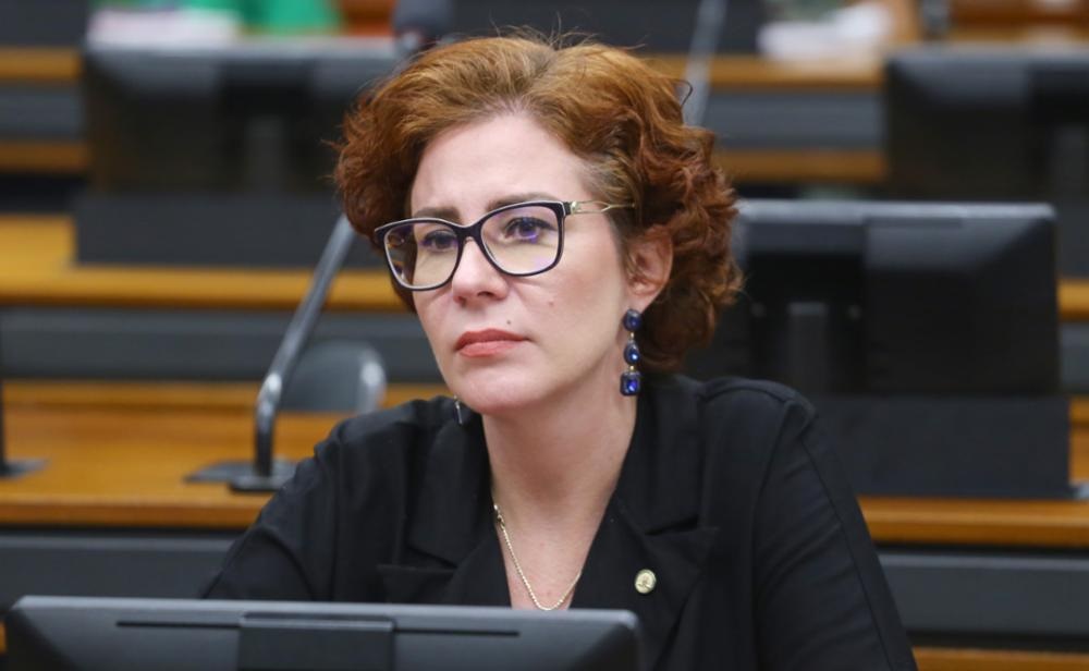 A deputada licenciada Carla Zambelli (PL-SP) séria, de óculos, roupa preta e cabelo solto, sem olhar para a câmera