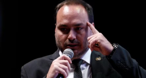 O vereador Carlos Bolsonaro (PL-RJ) sério, falando em microfone, com uma das mãos na cabeça