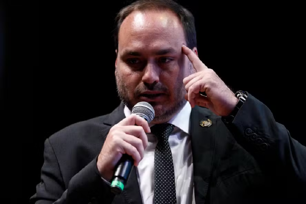 O vereador Carlos Bolsonaro (PL-RJ) sério, falando em microfone, com uma das mãos na cabeça