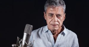 Chico Buarque olhando sério para a câmera, perto de microfone, de camisa social clara