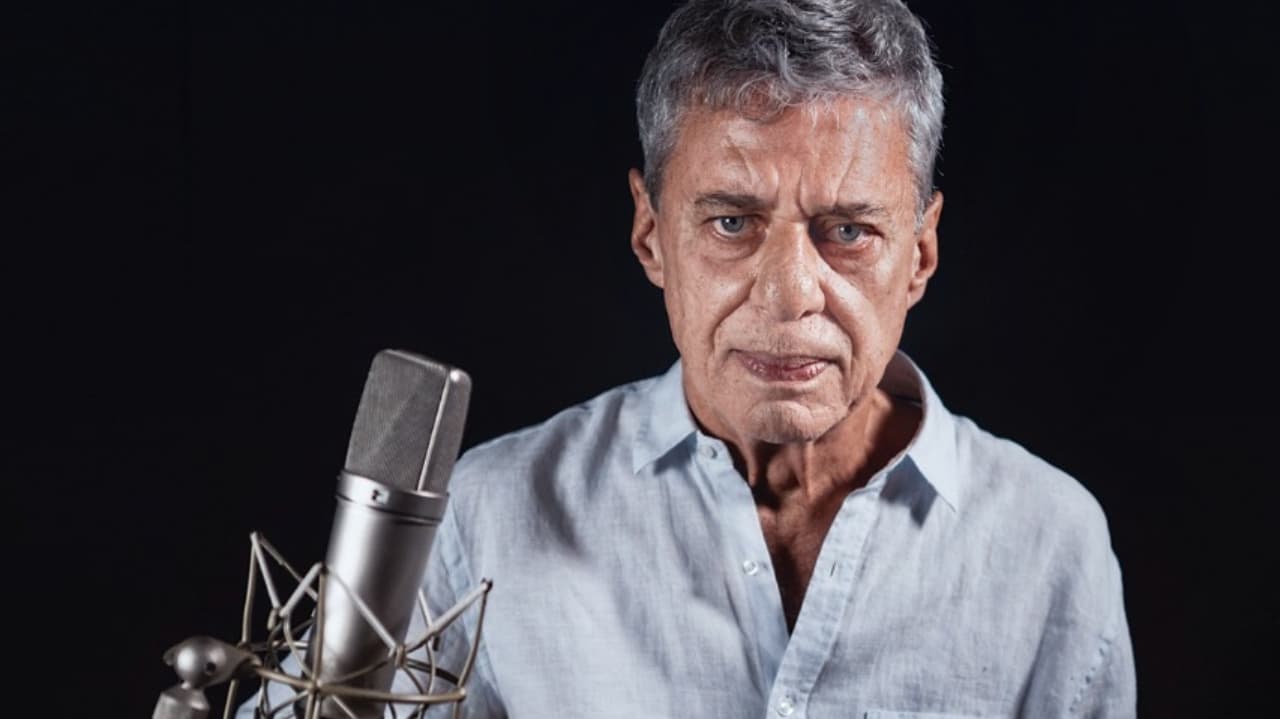 Chico Buarque olhando sério para a câmera, perto de microfone, de camisa social clara