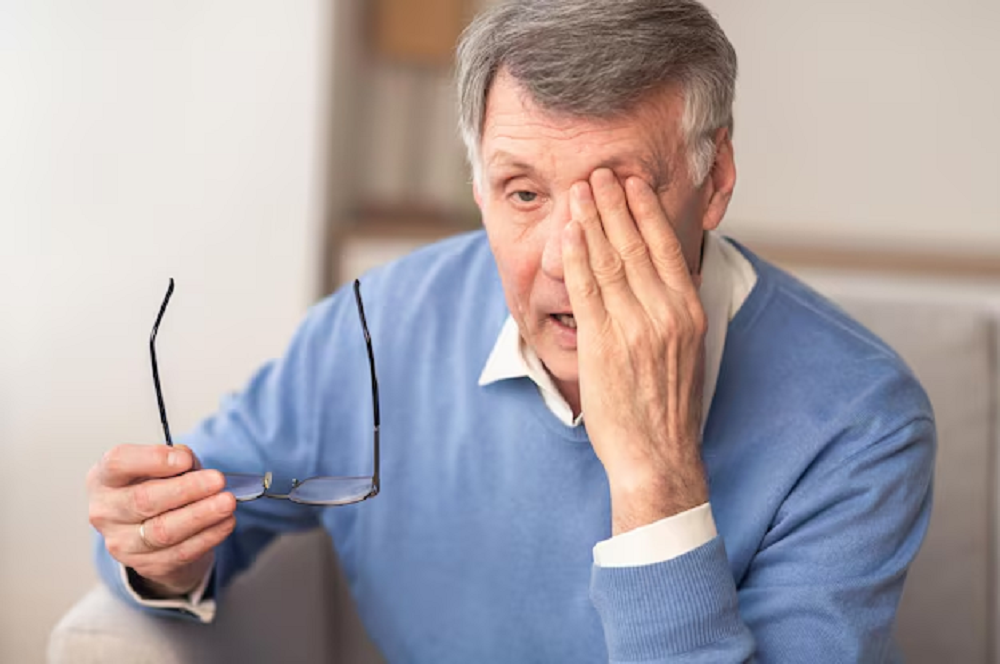 Idoso de roupa azul com mão no rosto, segurando óculos