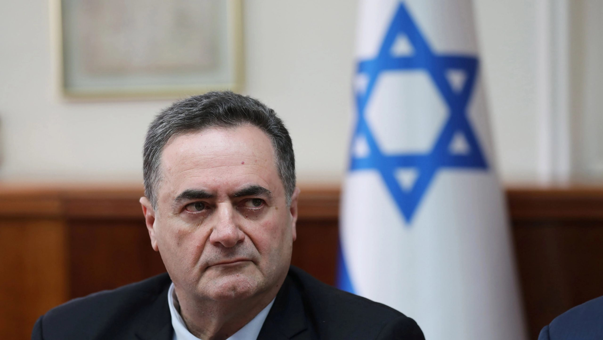 Ministro da Defesa, Israel Katz, sério, olhando para o lado esquerdo, com bandeira de Israel ao fundo