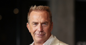 Kevin Costner sério, de roupa bege, sem olhar para a câmera