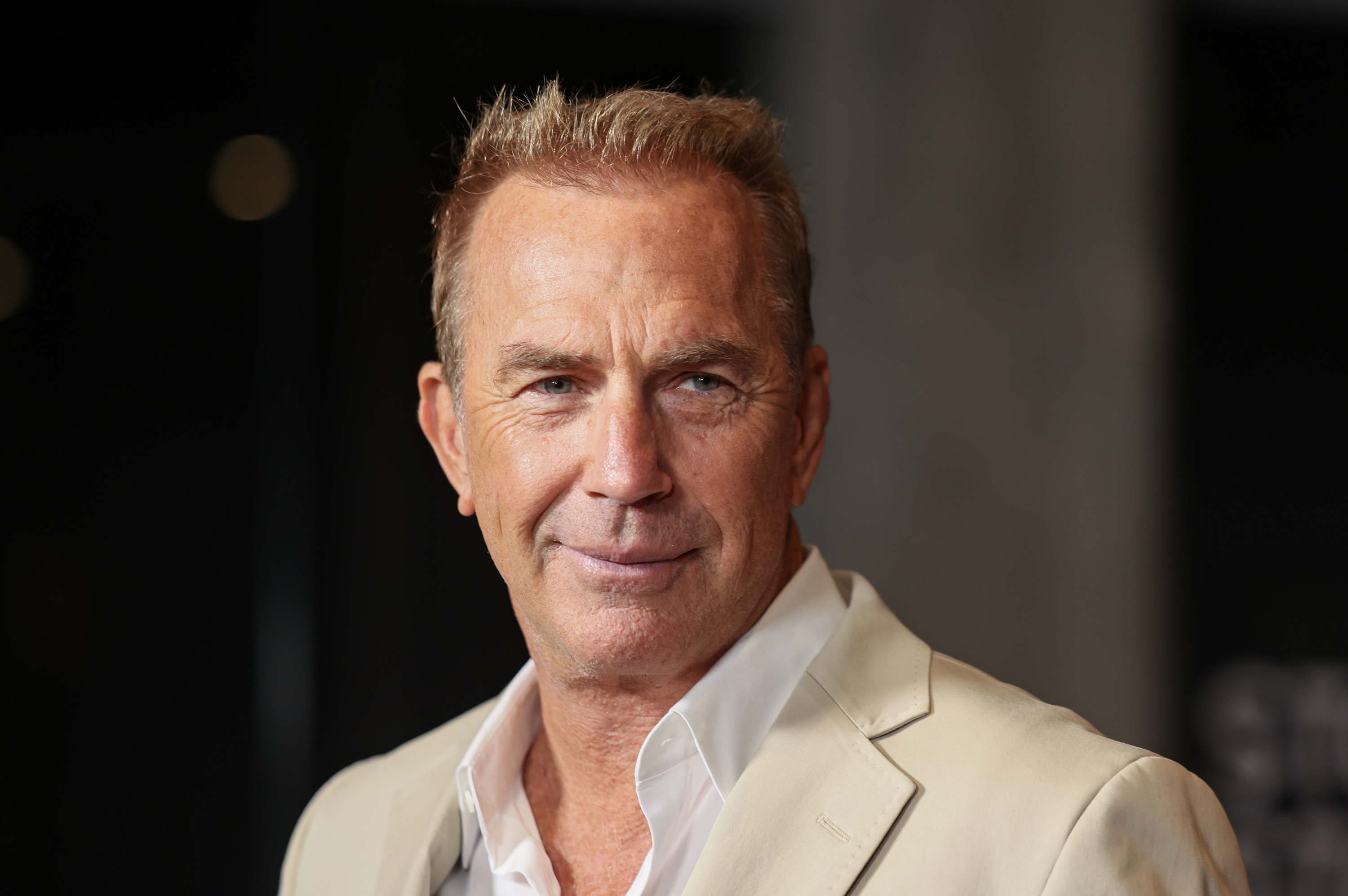 Kevin Costner sério, de roupa bege, sem olhar para a câmera