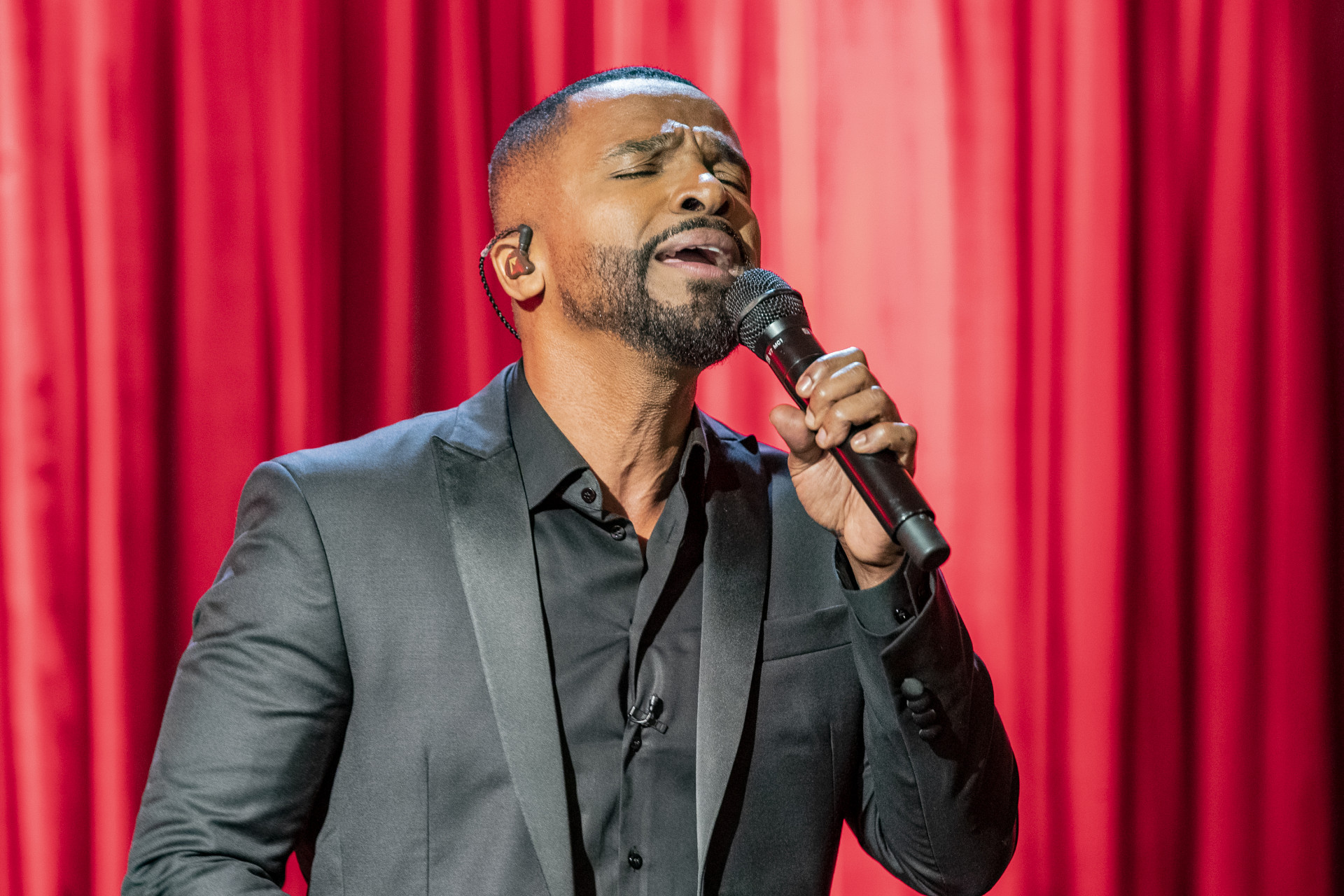 cantor Alexandre Pires cantando em microfone, de olhos fechados, com roupa social preta