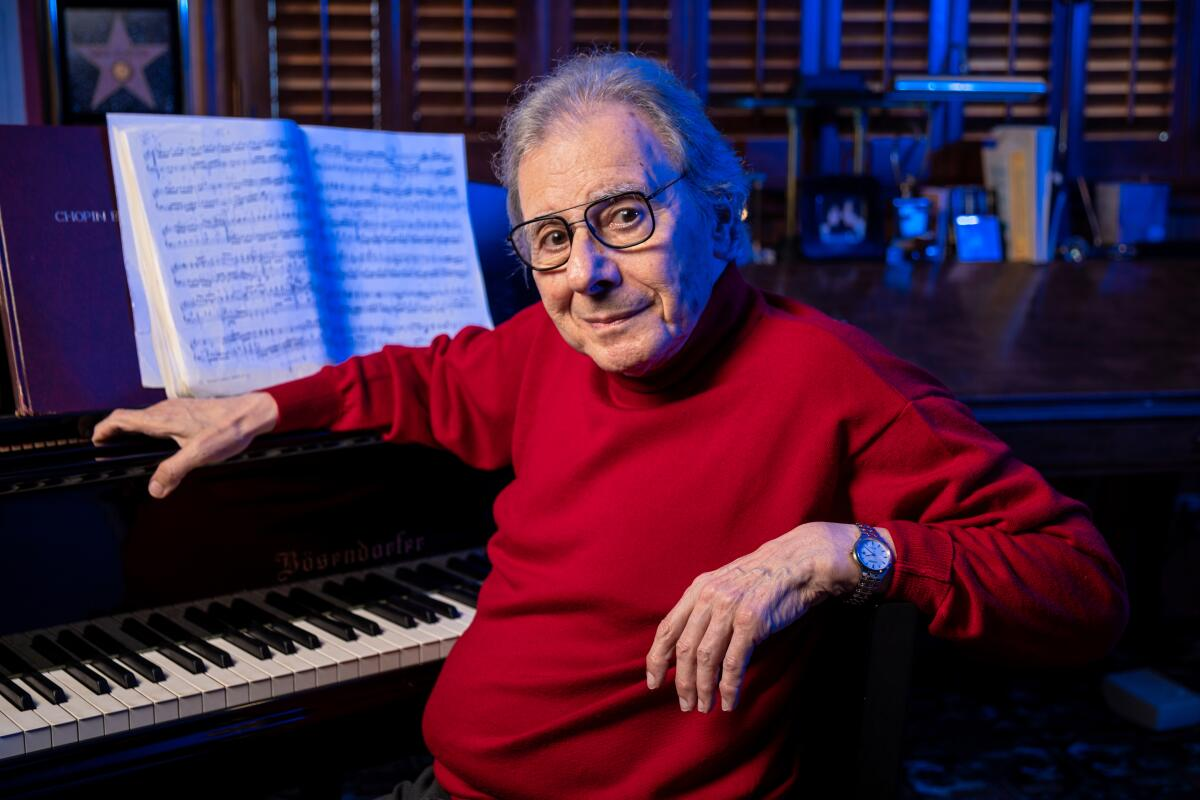 Lalo Schifrin de roupa vermelha e óculos de grau, sentado perto de piano, sério