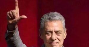 Chico Buarque fazendo um L com os dedos e sorrindo de maneira tímida, com um dos braços levantados