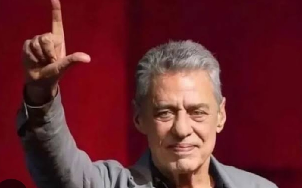 Chico Buarque fazendo um L com os dedos e sorrindo de maneira tímida, com um dos braços levantados