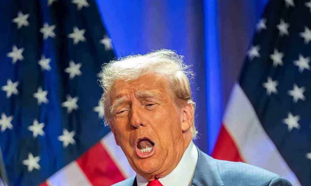 O presidente dos Estados Unidos, Donald Trump, falando com expressão séria, com bandeiras dos EUA no fundo