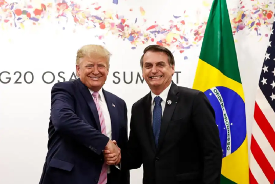 Donald Trump e Jair Bolsonaro em aperto de mãos, sorrindo para a câmera