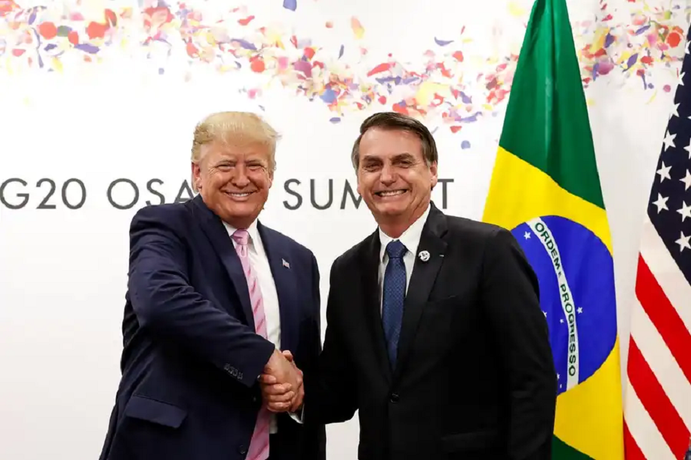 Donald Trump e Jair Bolsonaro em aperto de mãos, sorrindo para a câmera