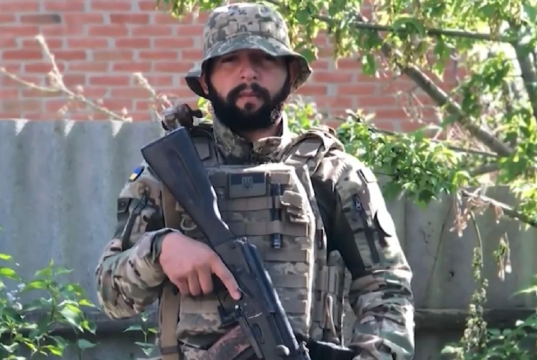 O brasileiro Rafael Paixão fardado, segurando arma, olhando sério para a câmera