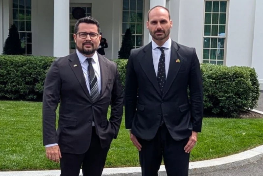 O jornalista Paulo Figueiredo e o deputado federal licenciado Eduardo Bolsonaro (PL) em pé lado a lado, olhando para a câmera