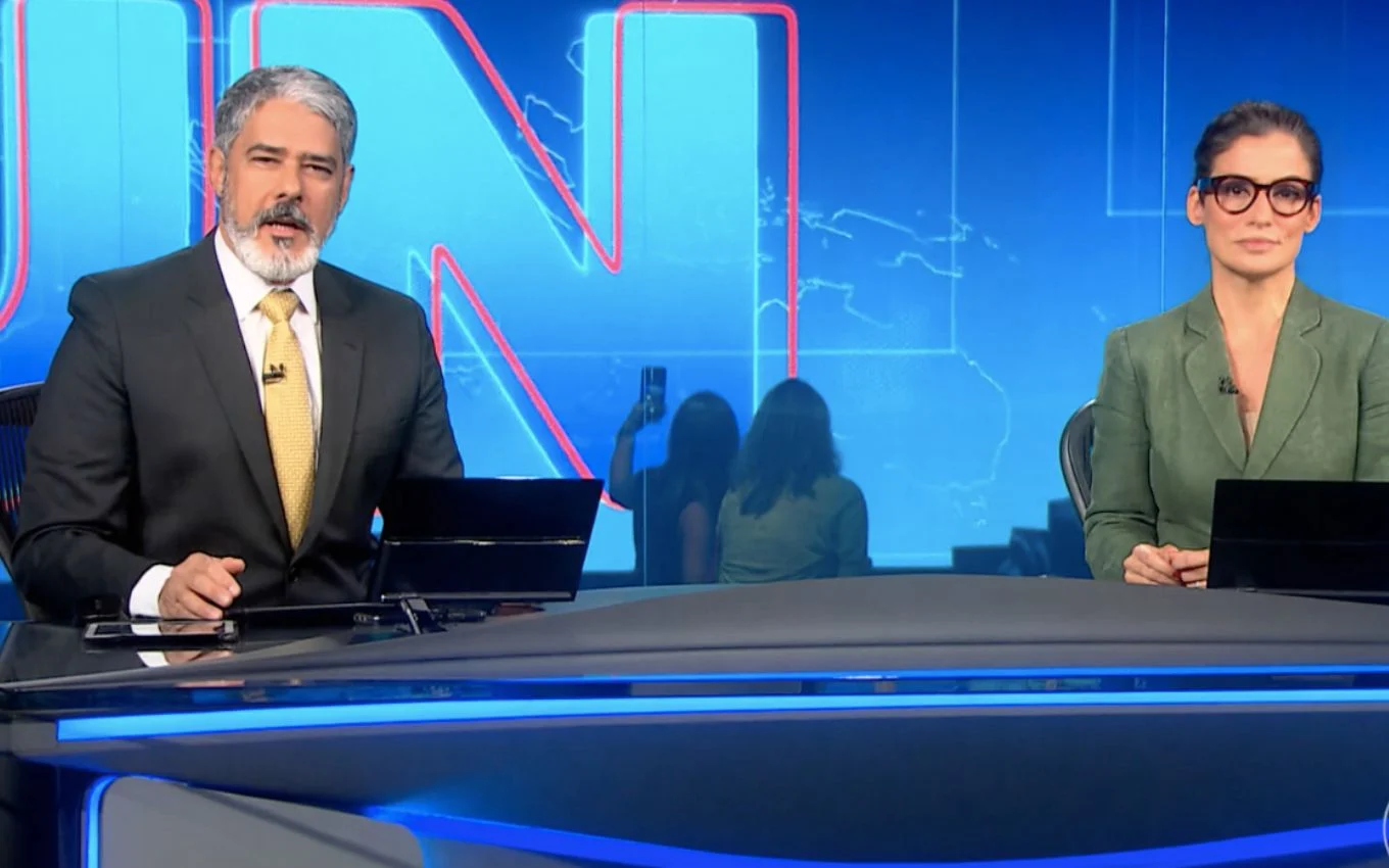 William Bonner e Renata Vasconcellos na bancada do Jornal Nacional, sérios, olhando para a câmera