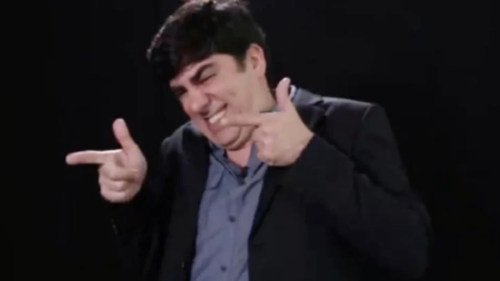O humorista Marcelo Adnet rindo e fazendo arminha com as mãos, sem olhar para a câmera