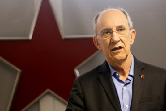 Rui Falcão falando, sério, com estrela vermelha ao fundo