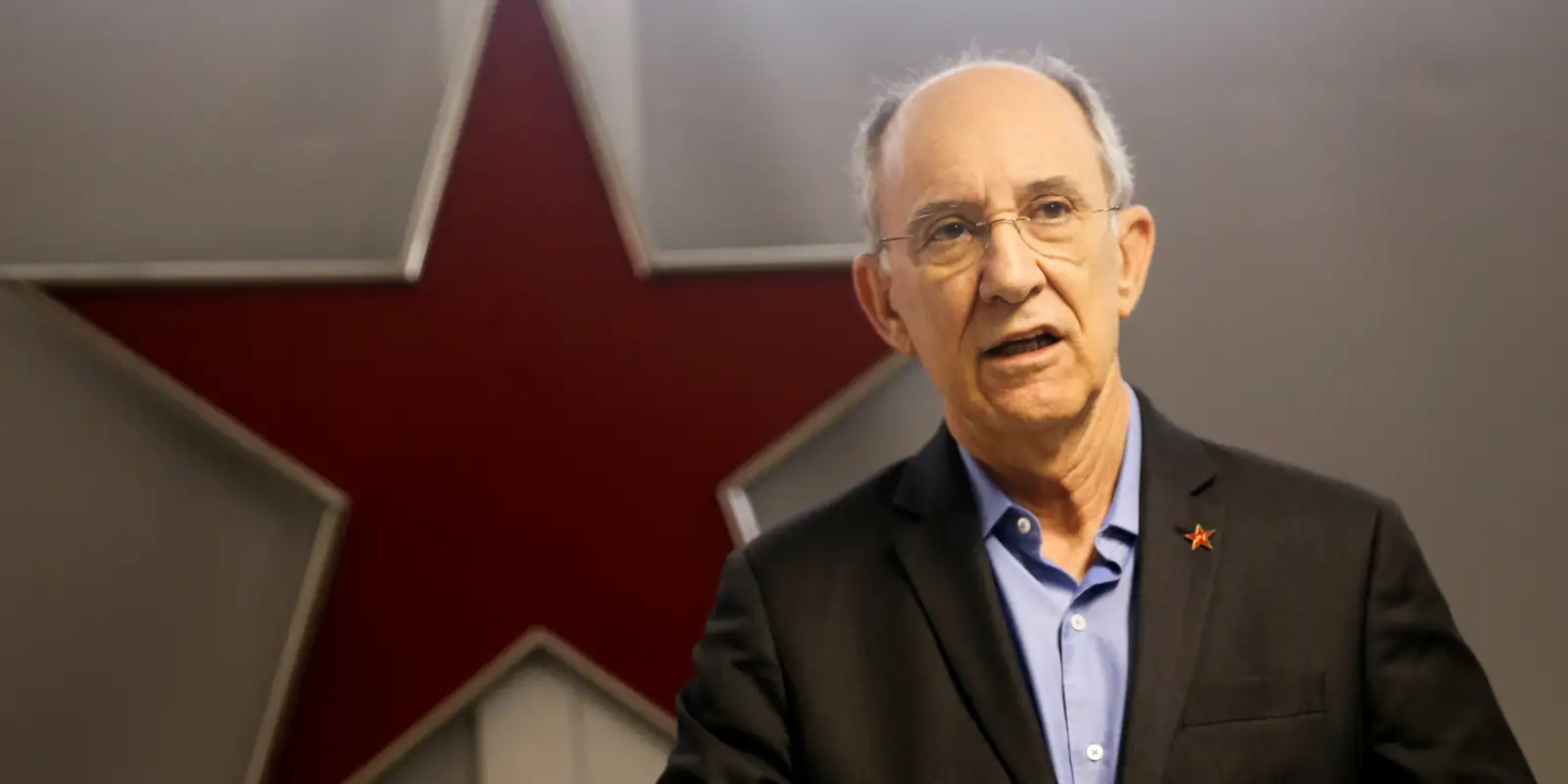 Rui Falcão falando, sério, com estrela vermelha ao fundo