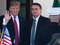 Donald Trump acenando e Jair Bolsonaro sério, ambos em pé, lado a lado
