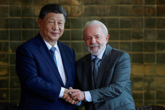 O presidente Xi Jinping e o presidente Lula sorrindo e olhando para a câmera durante aperto de mãos