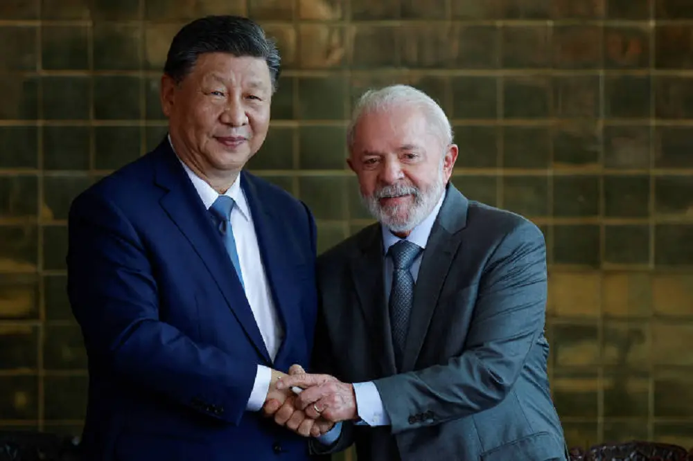 O presidente Xi Jinping e o presidente Lula sorrindo e olhando para a câmera durante aperto de mãos