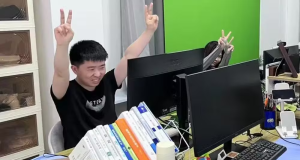 Shui Zhou em um escritório, sorrindo em frente ao computador, com os braços erguidos
