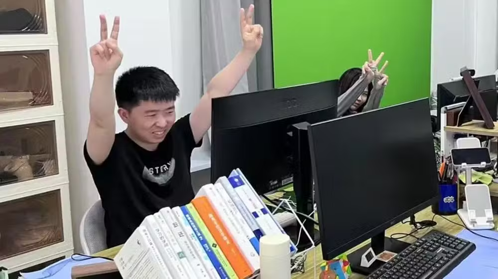 Shui Zhou em um escritório, sorrindo em frente ao computador, com os braços erguidos