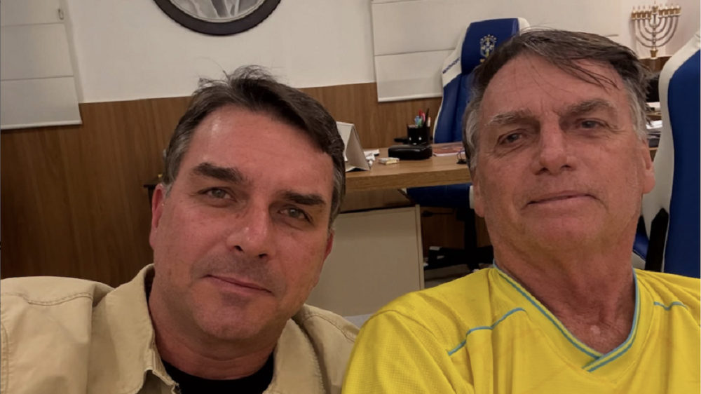 O senador Flávio Bolsonaro (PL-RJ) e Jair Bolsonaro posando para selfie lado a lado, sérios