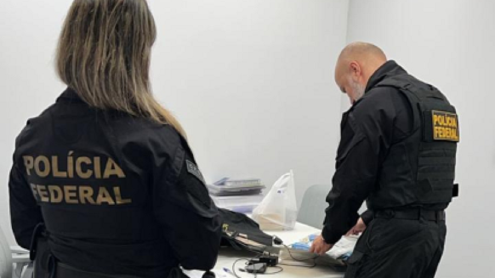 Agentes da Polícia Federal em pé, de costas, mexendo em objetos que estão em cima de mesa