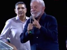 O presidente Lula (PT) de roupa social escura, falando em microfone e apontando, sério