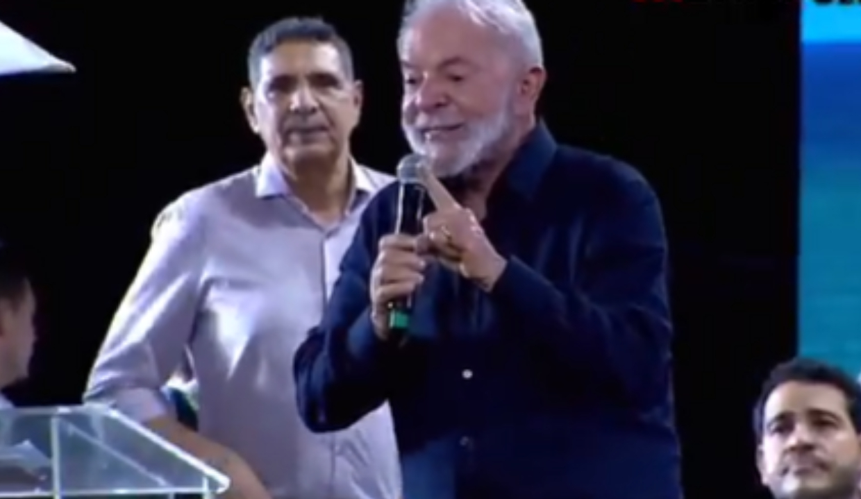 O presidente Lula (PT) de roupa social escura, falando em microfone e apontando, sério