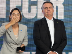 A ex-primeira-dama Michelle Bolsonaro (PL) e o ex-presidente Jair Bolsonaro (PL) lado a lado, em pé, ele sério e ela mandando beijo