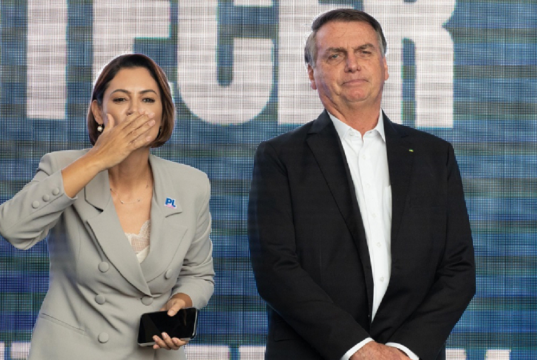 A ex-primeira-dama Michelle Bolsonaro (PL) e o ex-presidente Jair Bolsonaro (PL) lado a lado, em pé, ele sério e ela mandando beijo