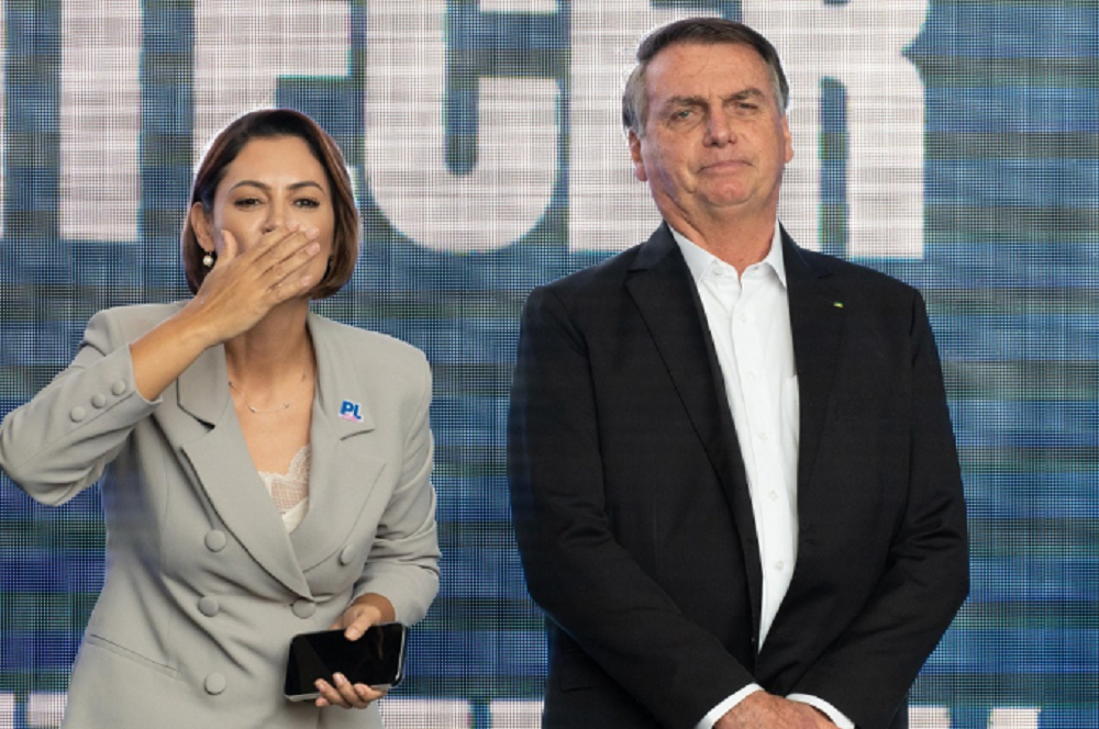 A ex-primeira-dama Michelle Bolsonaro (PL) e o ex-presidente Jair Bolsonaro (PL) lado a lado, em pé, ele sério e ela mandando beijo