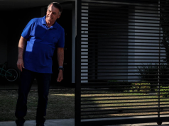 Jair Bolsonaro (PL) de calça preta e camisa azul em pé perto de portão