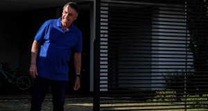 Jair Bolsonaro (PL) de calça preta e camisa azul em pé perto de portão