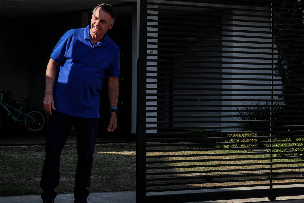 Jair Bolsonaro (PL) de calça preta e camisa azul em pé perto de portão