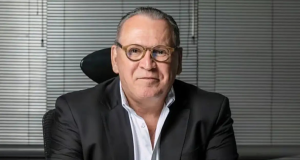 Sidney Oliveira, dono da Ultrafarma, sorrindo para a câmera, sentado, de óculos de grau e roupa social