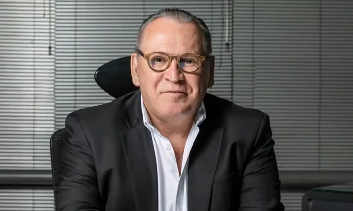 Sidney Oliveira, dono da Ultrafarma, sorrindo para a câmera, sentado, de óculos de grau e roupa social