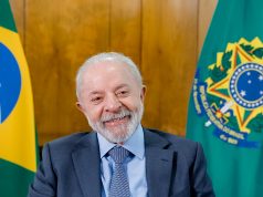 Presidente da República, Luiz Inácio Lula da Silva, sorrindo sem olhar para a câmera, com bandeiras brasileiras ao fundo