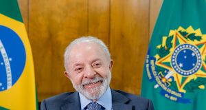 Presidente da República, Luiz Inácio Lula da Silva, sorrindo sem olhar para a câmera, com bandeiras brasileiras ao fundo