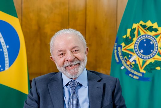 Presidente da República, Luiz Inácio Lula da Silva, sorrindo sem olhar para a câmera, com bandeiras brasileiras ao fundo
