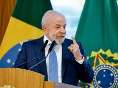 Lula sorrindo e apontando pra cima, sem olhar para a câmera, em pé, com bandeira do Brasil ao fundo