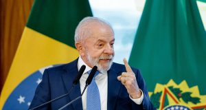Lula sorrindo e apontando pra cima, sem olhar para a câmera, em pé, com bandeira do Brasil ao fundo