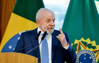 Lula sorrindo e apontando pra cima, sem olhar para a câmera, em pé, com bandeira do Brasil ao fundo