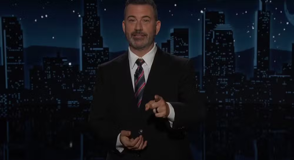 Jimmy Kimmel de roupa social, em pé, falando e apontando para a câmera