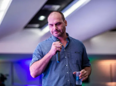 Eduardo Bolsonaro falando em microfone, cabisbaixo, em pé, de camisa social azul, segurando copo de água