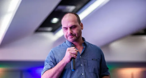 Eduardo Bolsonaro falando em microfone, cabisbaixo, em pé, de camisa social azul, segurando copo de água