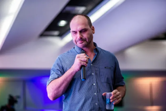 Eduardo Bolsonaro falando em microfone, cabisbaixo, em pé, de camisa social azul, segurando copo de água